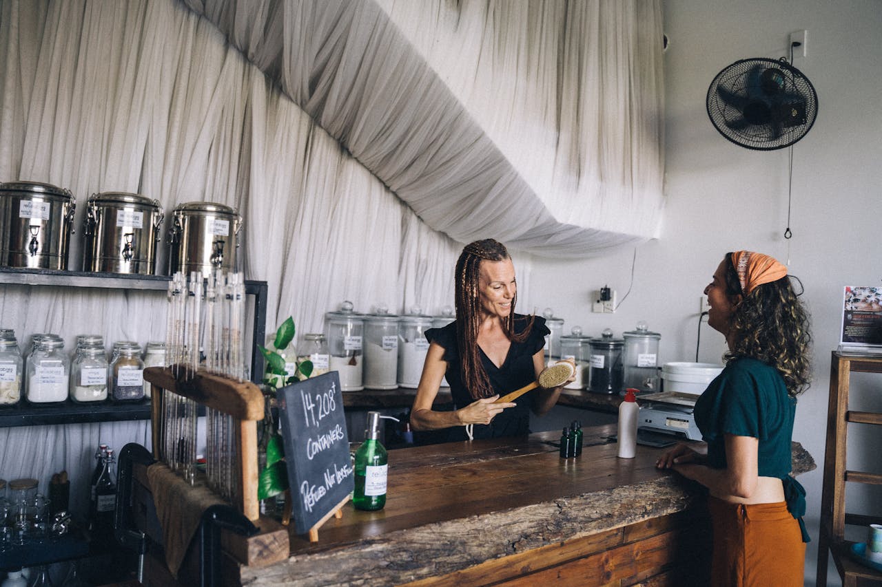 why-choose-us Two women engaging at an eco-friendly shop counter with refillable containers and glass jars.